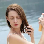 Жена пръска спрей върху косата си на плажа – грижа за косата през лятото и защита от слънцето