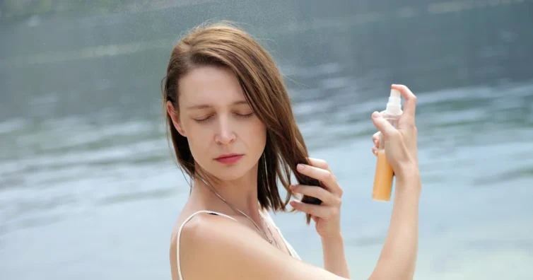 Жена пръска спрей върху косата си на плажа – грижа за косата през лятото и защита от слънцето